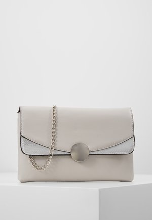 Petit sac pochette gris clair avec bandoulière chaîne argentée et fermoir rond argenté sur une surface blanche contre un fond clair uni.