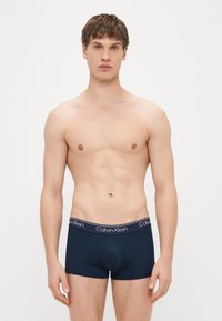 Tumši zilas bokseršorti no gludas auduma ar elastīgu jostu, uz kuras ir redzams logotips. Piegriezums cieši pieguļošs, dizains minimālistisks.