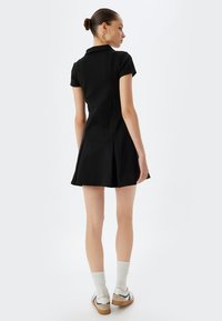 Vestido negro ajustado con cuello polo y mangas cortas, con una falda acampanada y detalles con pliegues. Combinado con calcetines blancos y zapatillas.