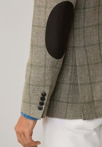 Blazer a motivo pied de poule grigio e beige con toppe nere sui gomiti. Presenta quattro bottoni neri sui polsini e una camicia di denim blu sotto.