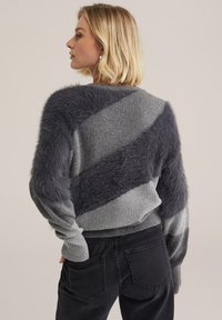 Grauer Strickpullover mit diagonaler Streifenoptik und weichen, flauschigen Akzenten, kombiniert mit schwarzen Jeans. Strukturierter Stoff und gerippte Bündchen.