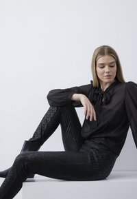 Zwarte doorschijnende blouse met een strik om de hals, gecombineerd met zwarte textuurleggings met een kantpatroon. Model poseert zittend.