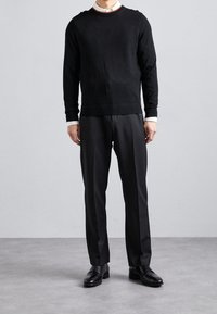 Homme portant un pull noir, une chemise blanche à col, un pantalon de costume noir et des chaussures de ville noires, debout sur un sol gris devant un mur clair uni.