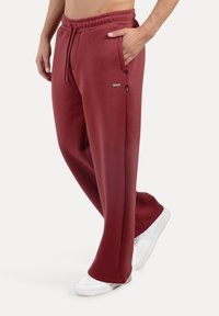 Burgunderfarvede joggingbukser lavet af blødt stof med elastisk talje, sidelommer og afslappet pasform, kombineret med hvide sneakers.