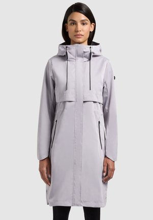 Woman wearing a light gray knee-length raincoat with a hood, front zipper, and zippered side pockets, standing against a plain background.