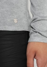 Szary sweter z dzianiny z ribbowym wykończeniem na dole i mankietach, z metalicznym logo w kolorze srebrnym umieszczonym blisko dołu. Czarne teksturowane spodnie są częściowo widoczne.