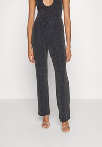 Svart, glittrande jumpsuit med djup V-ringning och åtsittande midja. Utsvängda ben sträcker sig ner till anklarna, parat med bandtofflor.