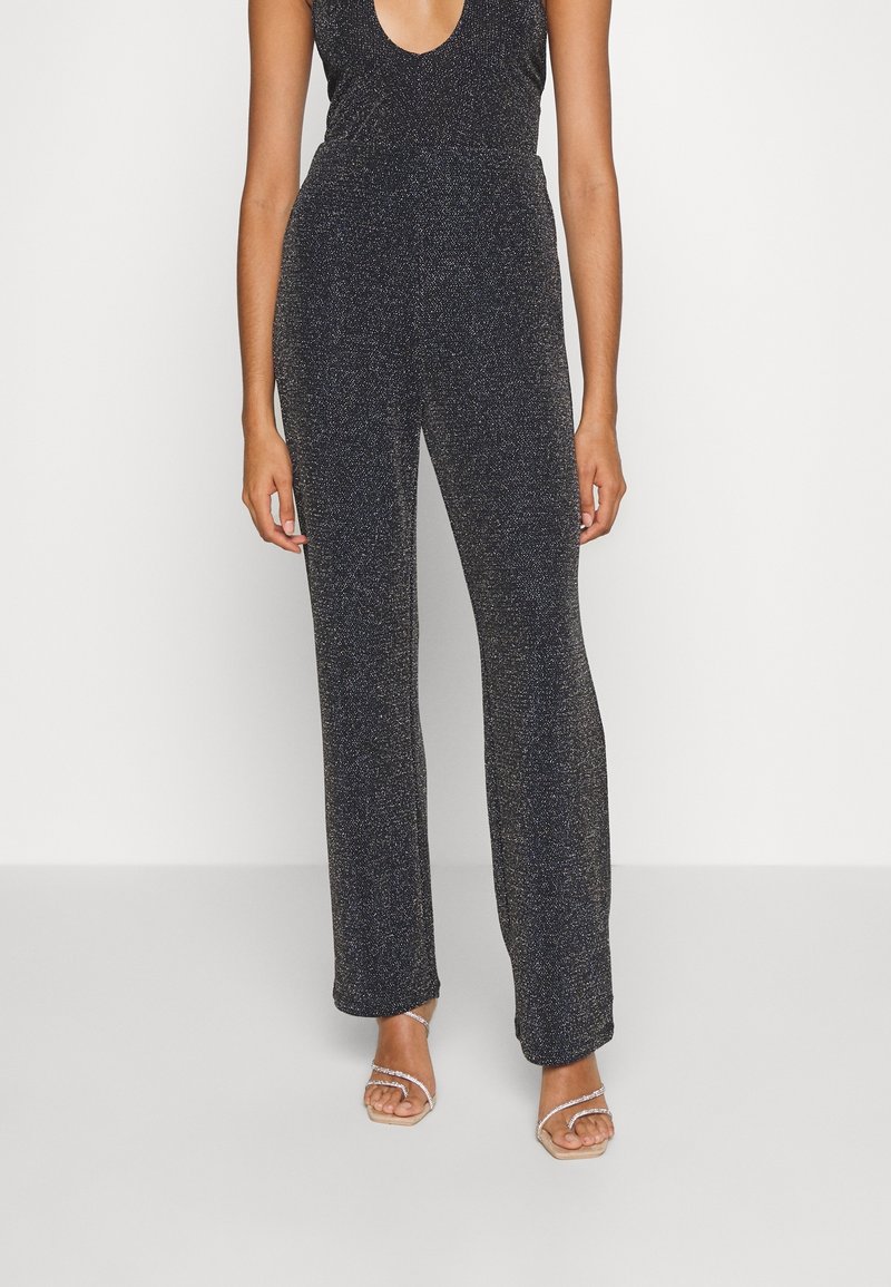 Svart, glittrande jumpsuit med djup V-ringning och åtsittande midja. Utsvängda ben sträcker sig ner till anklarna, parat med bandtofflor.