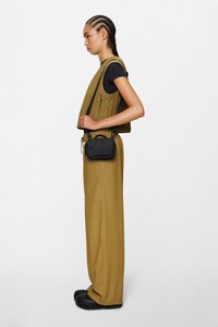 Chaleco acolchado verde oliva sobre una camiseta negra, combinado con pantalones anchos beige y zapatos negros, y un bolso pequeño negro de bandolera.