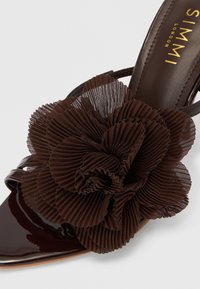 Smeđi lakirani otvoreni potpetica s velikim, teksturiranim smeđim naboranim cvjetnim ukrasom. Cipela ima tanke remenčiće i glatku završnu obradu.