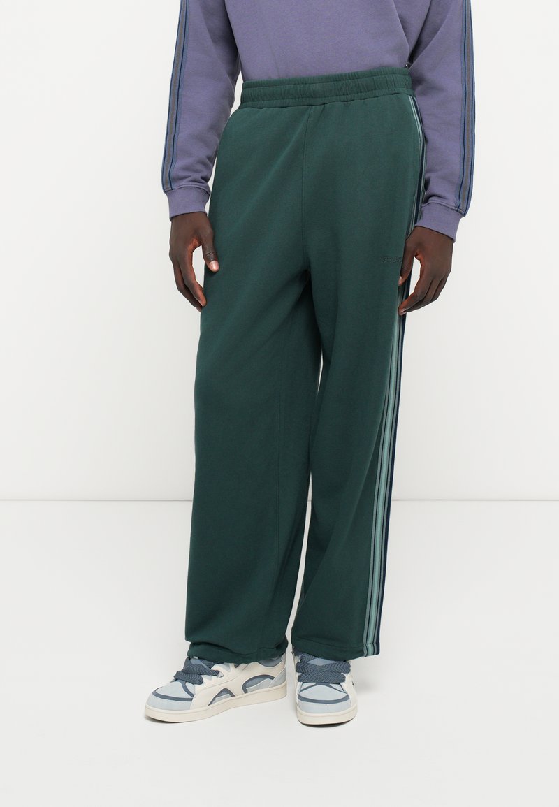 Green sweatpants with a relaxed fit, elastic waistband, side stripes, and soft texture. Paired with light blue sneakers.