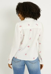 Blusa blanca texturizada con bordado floral en rosa, azul y verde. Cuenta con mangas largas, cuello con volantes y puños abotonados.