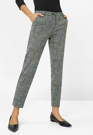 Vrouw draagt grijze patroon taps toelopende broek met voorzakken en zwarte puntige platte schoenen, staand tegen een witte achtergrond.