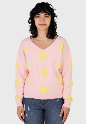 Giovane donna che indossa un maglione rosa con cuori gialli e jeans blu strappati, in piedi contro uno sfondo chiaro e semplice.