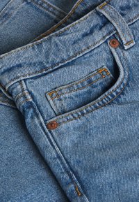 Närmre bild av en ficka på blå denimjeans med orange sömnad och kopparnitar på strukturerad tyg.