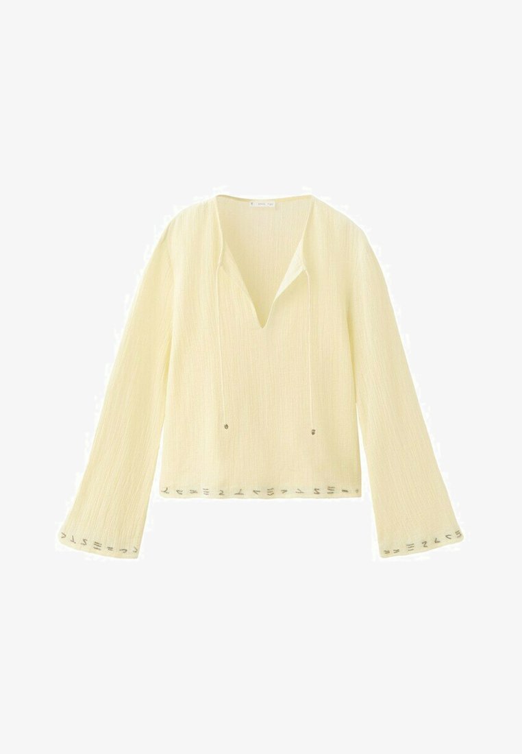 Lysegul bluse med lange ærmer, V-hals, løs pasform og små metalliske pynt langs ærmekanter og nederst på blusen.