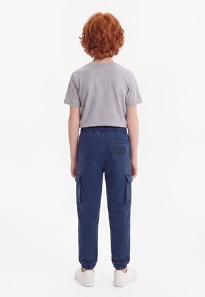 Enfant aux cheveux roux bouclés, dos tourné, portant un t-shirt gris à manches courtes, un pantalon cargo bleu marine et des baskets blanches.