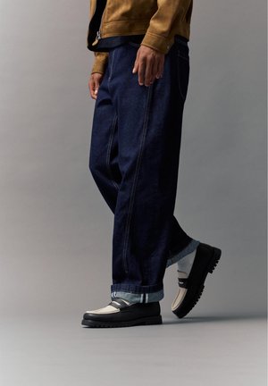 Person iført mørkeblå jeans med vidde ben, tanjakke og sort-hvide loafers, stående mod en ensfarvet grå baggrund.