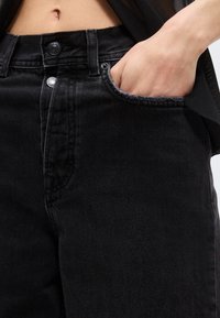 Άτομο που φοράει μαύρο τζιν ψηλής μέσης με κουμπιά, χέρι μερικώς μέσα στην μπροστινή τσέπη, δείχνοντας τη μέση και ανοιχτόχρωμο δέρμα.