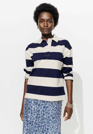 Polo à manches longues rayé bleu marine et blanc avec un col, associé à une jupe bleue ornée de sequins. Présente des manches retroussées et une patte de boutonnage devant.