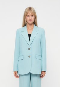 Woman with long blonde hair wearing a light blue tailored blazer with two buttons and matching trousers against a plain background.