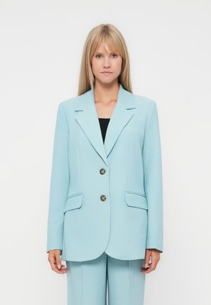 Kvinne med langt blondt hår iført en lys blå skreddersydd blazer med to knapper og matchende bukser foran en ensfarget bakgrunn.