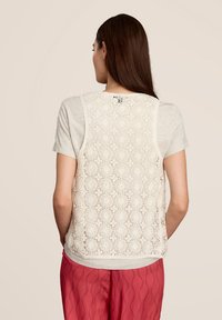 Top à dos en dentelle présentant des motifs floraux complexes en crème, avec un devant en coton gris doux et des manches courtes. Porté avec un pantalon texturé corail.