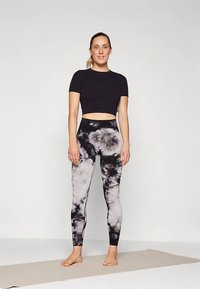 Nő áll mezítláb egy jógaszőnyegen fekete crop topot és fekete-fehér batikolt leggingset viselve, egyszínű háttér előtt.