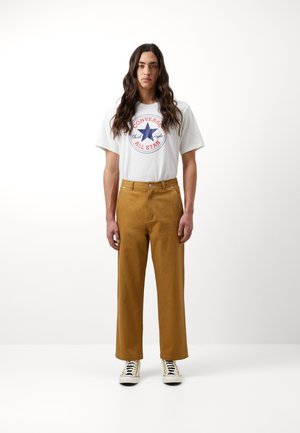 Camiseta blanca con logo de Converse en rojo y azul, combinada con pantalones anchos marrones y zapatillas a rayas en blanco y negro. Diseño sencillo.