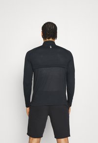 Long-sleeve black athletic shirt with a high collar, textured back panel, and flat seams. Paired with black shorts, features minimal design.