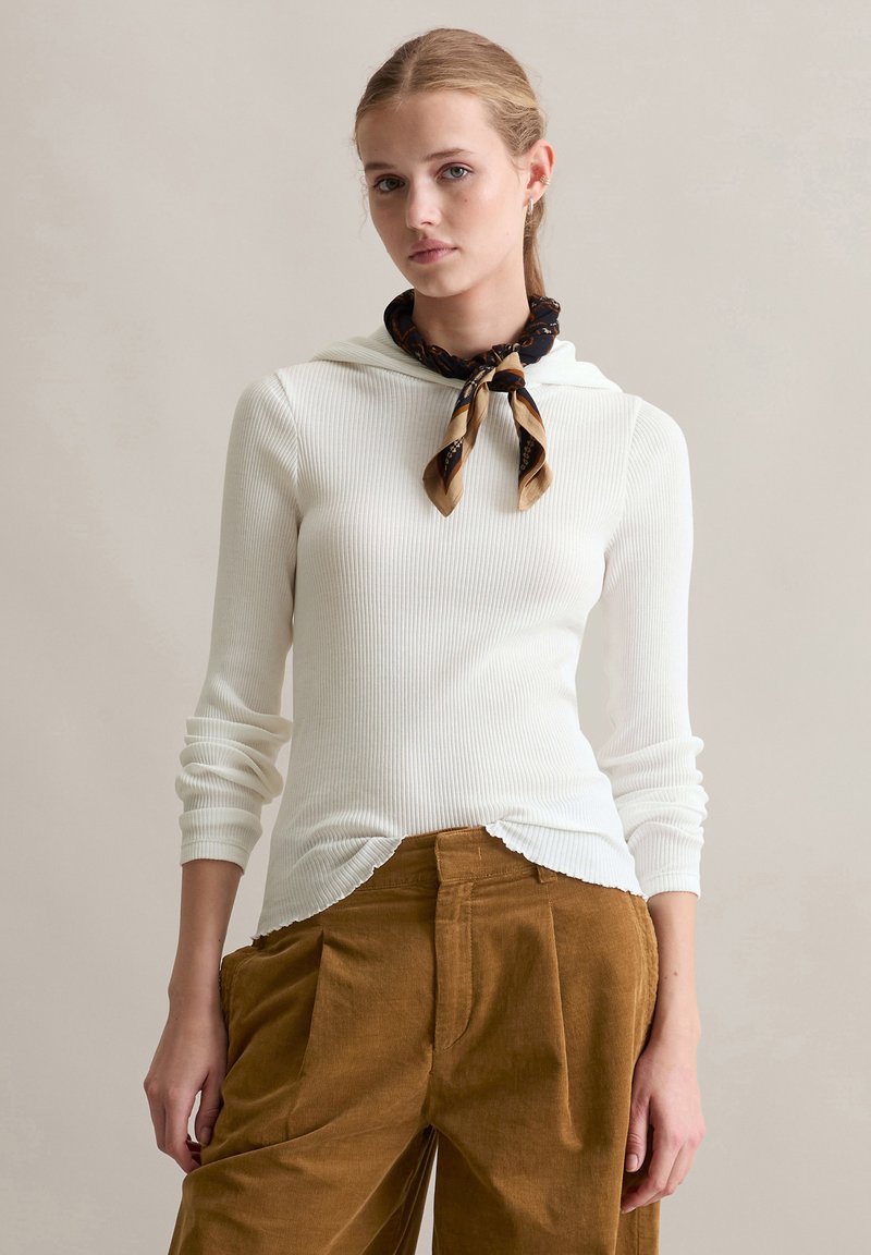 Geribbelde witte top met lange mouwen en een scalloped zoom, gecombineerd met bruine ribfluwelen hoge taillebroeken en een gestreepte halsdoek.