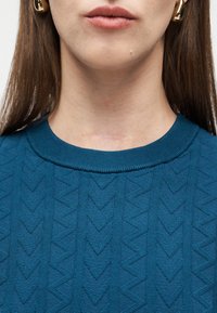 Κοντινό πλάνο του κάτω μέρους του προσώπου και του λαιμού μιας γυναίκας που δείχνει μια οριζόντια ουλή πάνω από τη λαιμόκοψη μιας μπλούζας με μπλε σχέδιο και χρυσά σκουλαρίκια κρίκους.