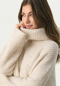 Beige strikket sweater med høj ribkant og struktureret mønster, med brede ærmer og et blødt, hyggeligt udseende.