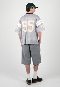 Hombre que lleva una camiseta deportiva de gran tamaño y estampado con el número 95, pantalones cortos a rayas hasta la rodilla, calcetines blancos, zapatos negros y una gorra de béisbol verde.