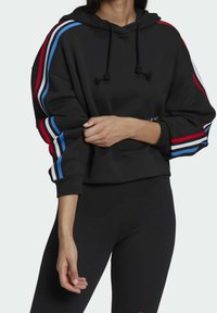 Svart hoodie i mjukt tyg, med röda, blå och vita randiga detaljer på ärmarna. Har en justerbar huva och ribbade mudd.