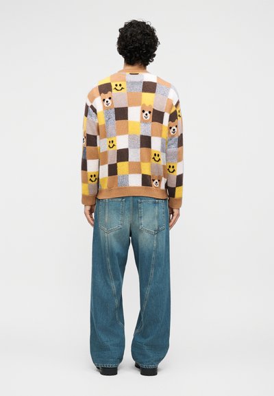 Άτομο με σγουρά μαλλιά φοράει πολύχρωμο πουλόβερ με καρό μοτίβα, πρόσωπα αρκούδας και χαμογελαστά σχέδια, συνδυασμένο με χαλαρό μπλε τζιν, στέκεται με την πλάτη γυρισμένη.