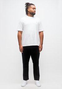 Άνδρας με dreadlocks, φορώντας λευκό T-shirt, μαύρο παντελόνι και λευκά αθλητικά παπούτσια, στέκεται πλάγια σε απλό φωτεινό φόντο.