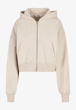 Haut de survêtement beige raccourci en tissu doux, avec une fermeture éclair à l'avant, deux grandes poches latérales et des poignets et un ourlet côtelés.