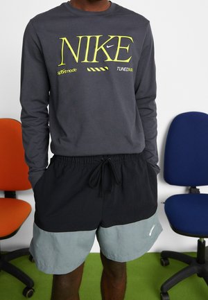 Langärmliges, dunkelgraues Nike-Shirt mit leuchtend gelber Schrift; kombiniert mit schwarzen und hellblauen Shorts mit einem Kordelzugbund.