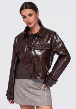 Femme portant une veste en cuir synthétique marron foncé brillante avec des boutons noirs et une mini-jupe à carreaux beige, posant avec une main sur la hanche.