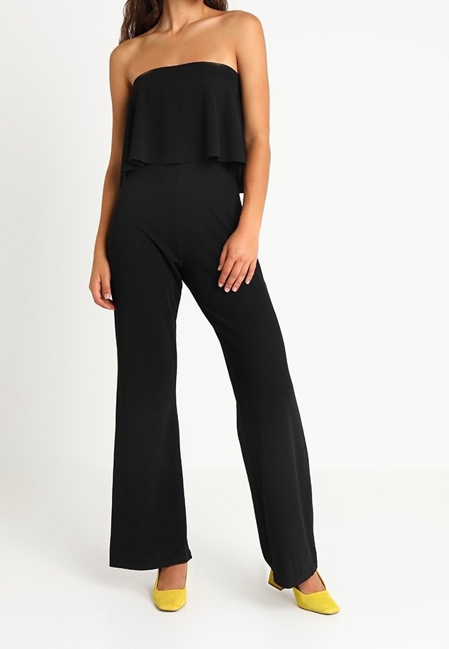 Femme portant une combinaison noire bustier à jambes larges et des chaussures à talons hauts jaunes, debout sur un fond blanc.