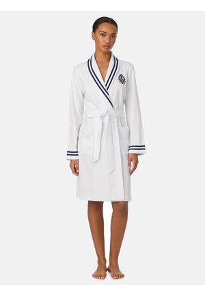 Donna in piedi scalza che indossa un accappatoio bianco fino al ginocchio con rifinitura blu navy e una cintura annodata in vita, con un piccolo stemma ricamato sul petto.