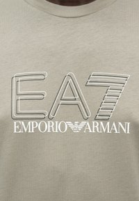 Λογότυπο EA7 με κείμενο και έμβλημα Emporio Armani σε μπεζ ύφασμα, τοποθετημένο στο κέντρο του στήθους ενός ατόμου που φοράει μπλουζάκι με στρογγυλή λαιμόκοψη.