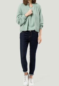 Cardigan lavorato a maglia verde chiaro sopra un top verde chiaro, abbinato a jeans blu scuro e sneakers bianche. Design semplice e aderente con polsini arrotolati.