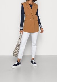 Brun dubbelknäppt ärmlös väst över en marinblå långärmad topp, kombinerat med vita raka jeans och marinblå sneakers, hållande en mönstrad väska.