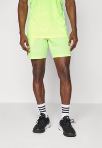 Haut et short de sport d'un vert néon vif, en tissu texturé, associés à des chaussures de sport noires et des chaussettes blanches rayées.