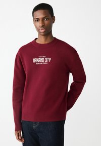 Bordowy sweter z prążkowanym okrągłym dekoltem i mankietami. Na przodzie znajduje się biały napis "TOKYO NAKANO CITY RENGA ZAKA". Materiał wydaje się gładki.