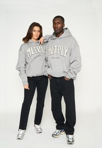 Grijze hoodies met de "MLTPLY" grafiek in dikke letters, gecombineerd met zwarte broeken en sportschoenen. Soepele textuur, relaxte pasvorm.