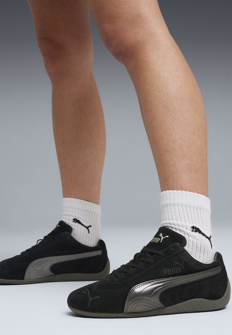 Jambes portant des chaussettes blanches Puma et des baskets en daim noir Puma avec des bandes de logo argentées sur une surface grise et un fond dégradé gris.