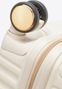 Walizka z twardą skorupą w kremowym kolorze, z złotym zamkiem błyskawicznym i szarymi akcentami na kółkach. Charakterystyczny okrągły detal metalowy na kole.